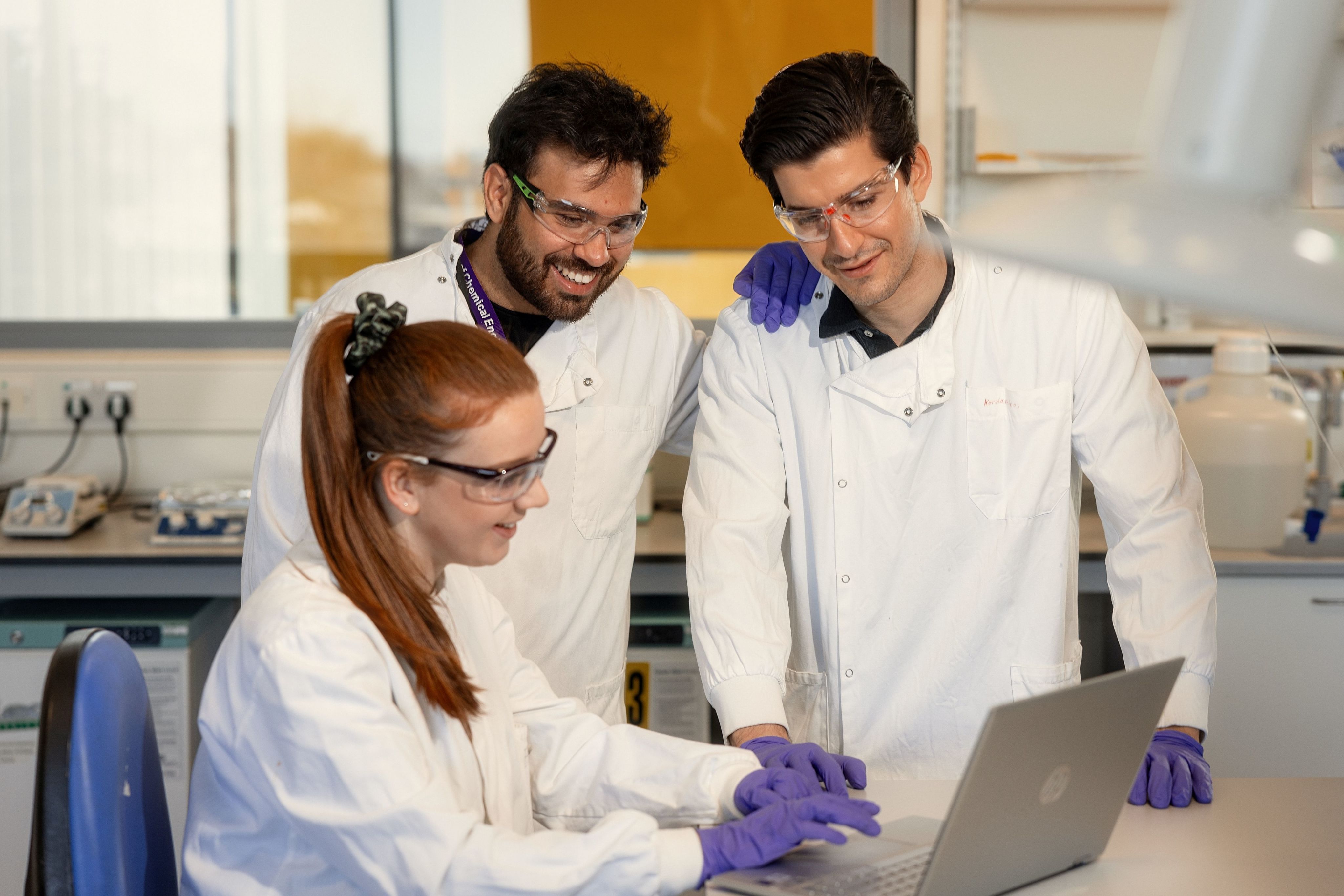 Three people in white lab coats looking studiously at a laptop, grinning.